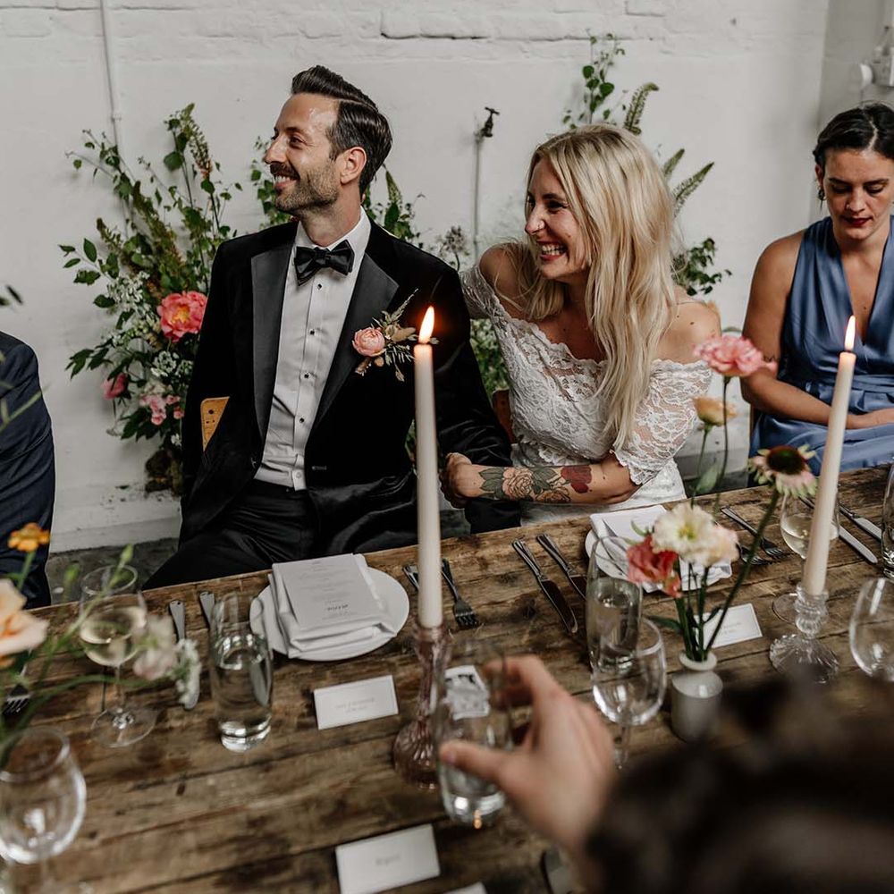 Bride in lace wedding top and groom in dark tux and bowtie laughing at wedding speeches in modern stylish reception room