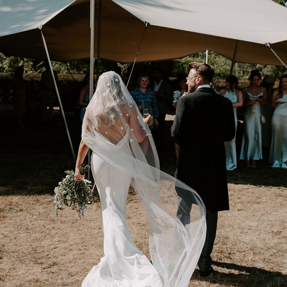 Bride in cowl back wedding dress with flower applique veil walking with the groom to the marquee reception 