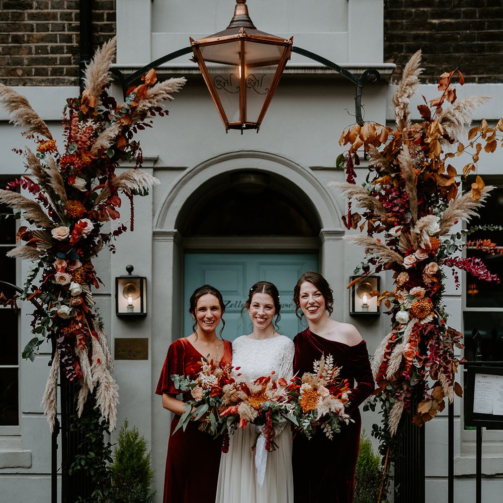 Bride in long sleeve lace vintage wedding dress and closed toe satin heels standing with bridesmaids in mismatched velvet bridesmaid dresses holding large autumnal bouquets standing under large floral arch with roses, peonies, foliage and pampas grass