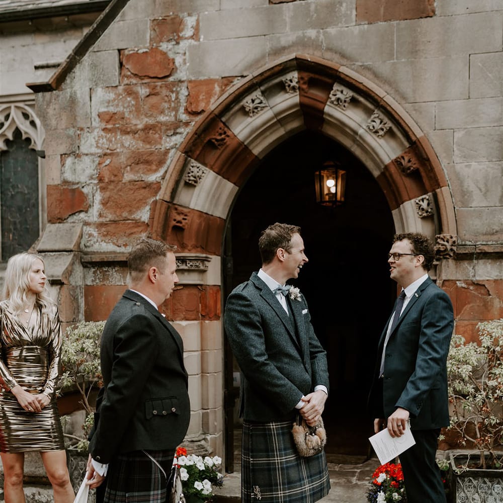 Groom and groomsmen stand outside the church as the guests arrive wearing kilts for Scottish traditional church wedding ceremony 
