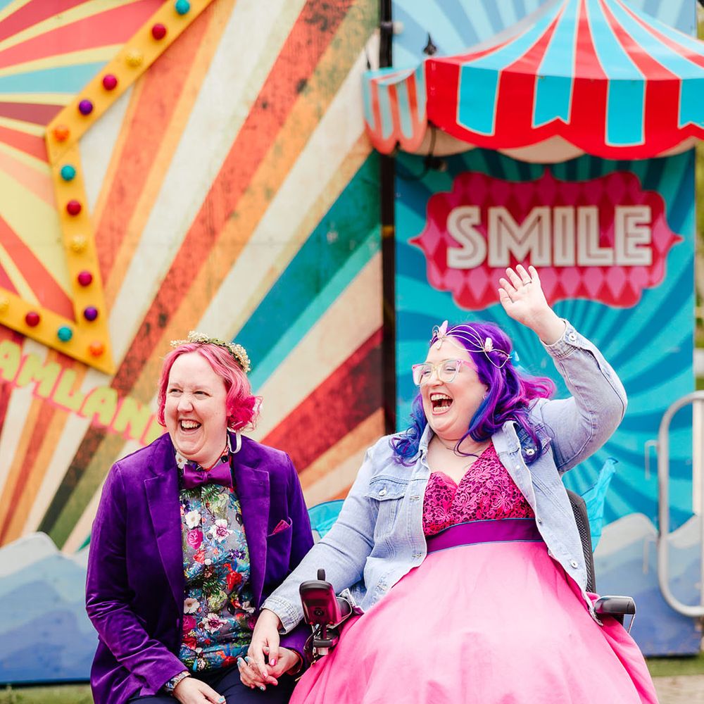Bride in wheelchair wearing personalised denim jacket, pink wedding dress and blue tights holding hands with bride in purple velvet blazer and black suit trousers with purple bowtie, pink patterned pocket square and colourful patterned shirt