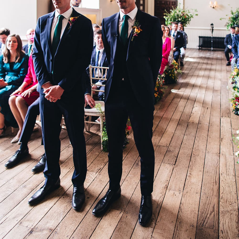 Groom stands in a three piece navy suit and green tie with colourful buttonhole at the altar with the best man also in a navy suit and green tie 
