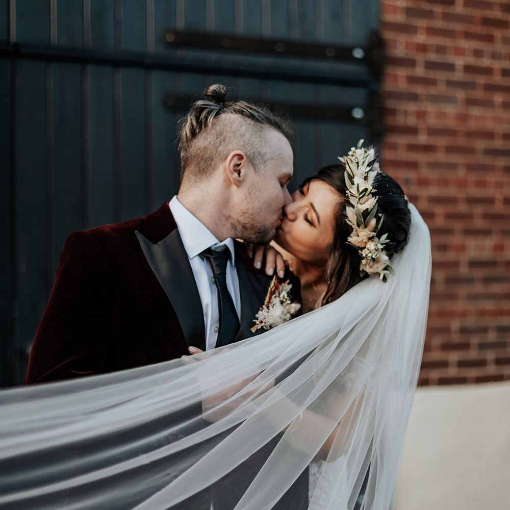 Groom in burgundy velvet suit jacket kissing the bride who wears a veil and boho dried flower crown 