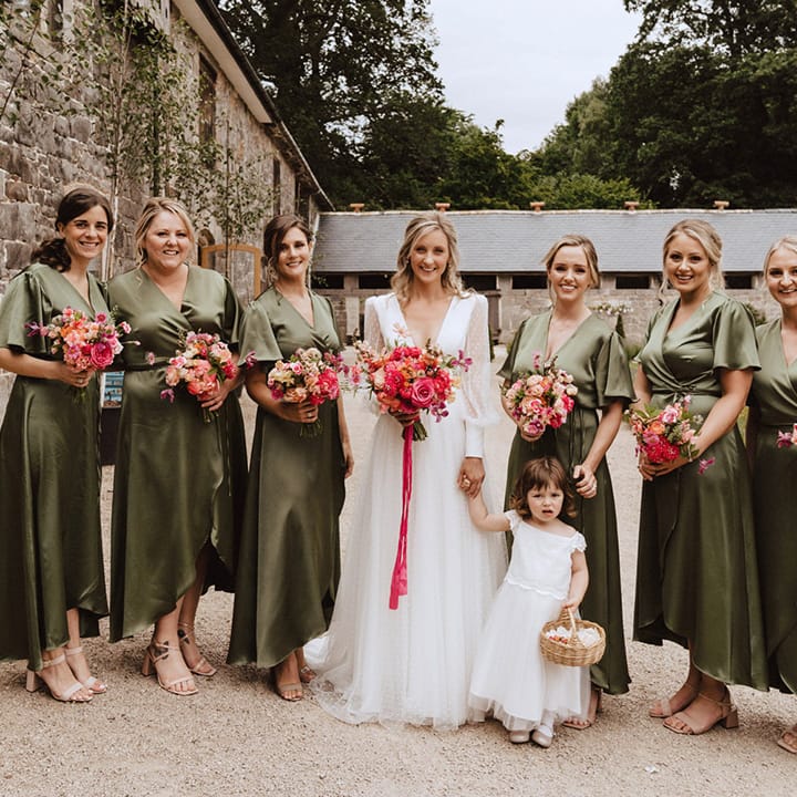Green satin wrap bridesmaid dresses from Rewritten Bridesmaids with the flower girl in a white dress holding hands with the bride in a Sassi Holford wedding dress all holding pink rose and peony bouquets