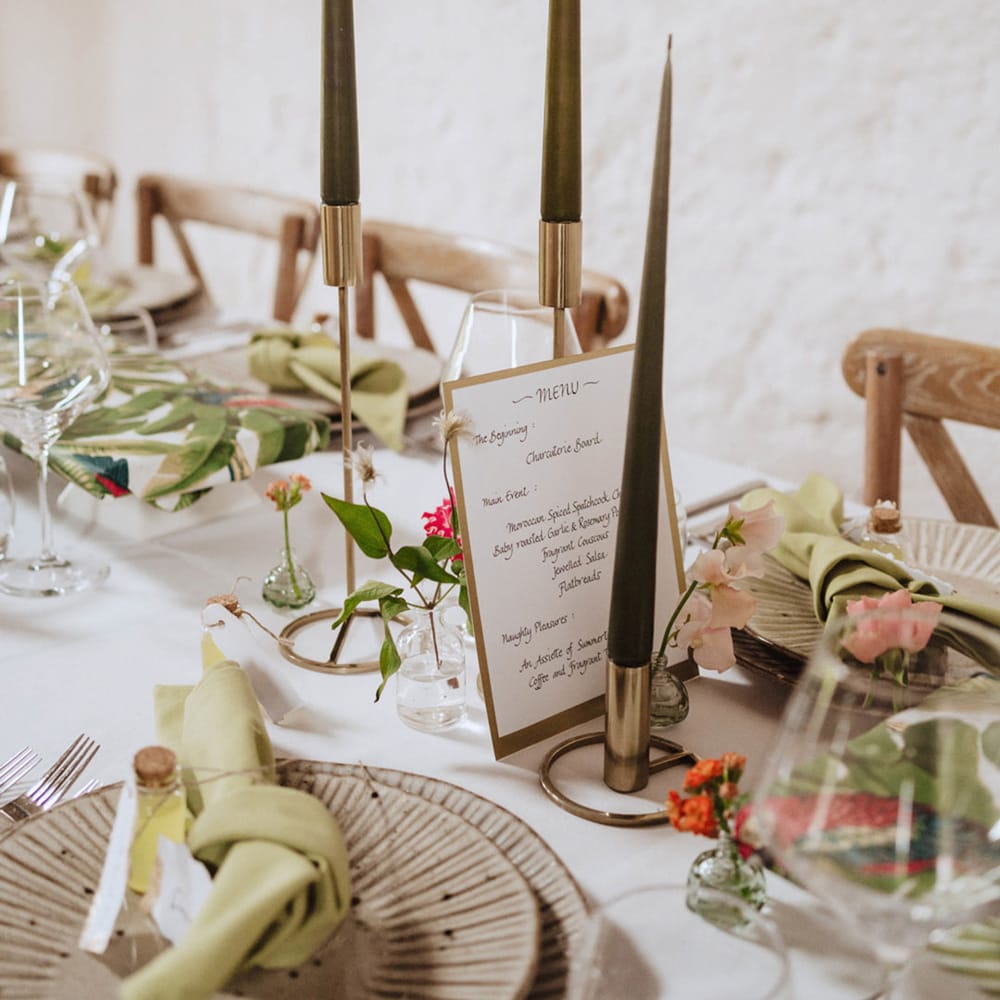 Dark green taper candles in gold candle holders with neutral coloured textured plates with green napkins and limoncello shots for favours 