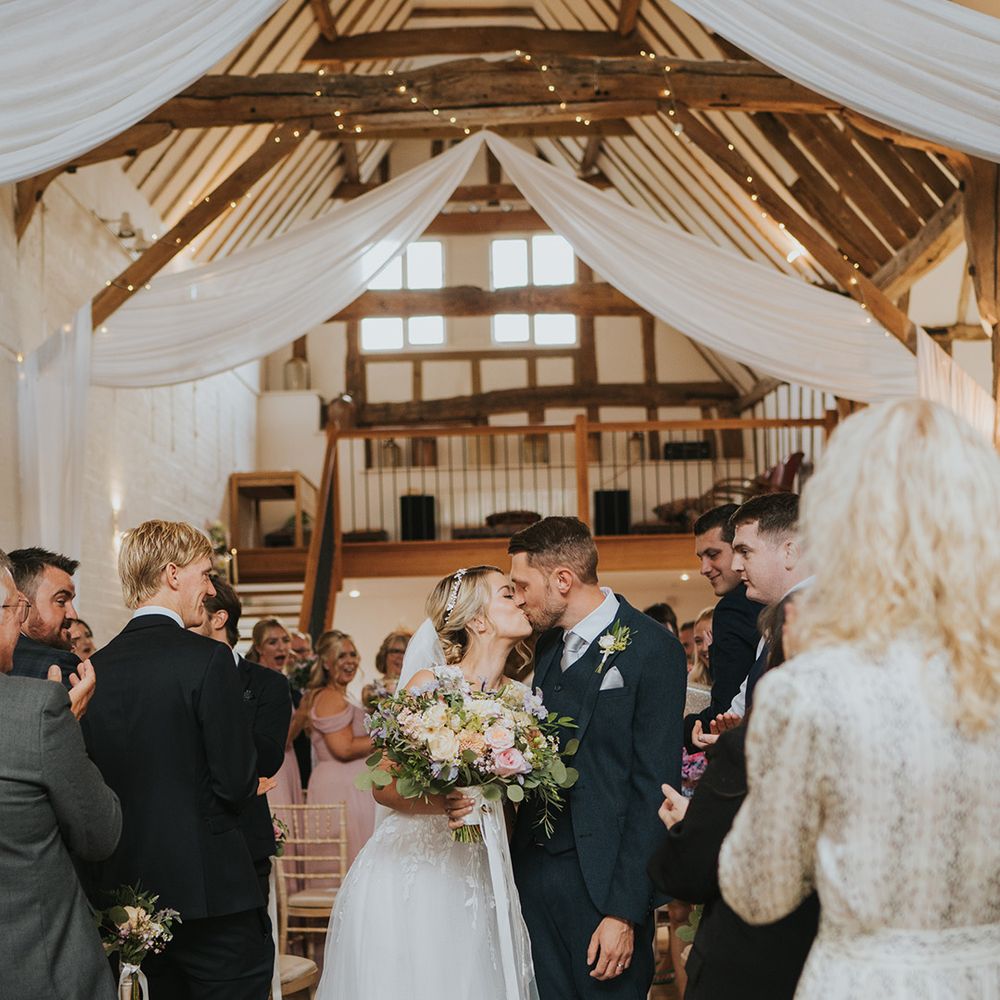 White drapery and fairy lights decorate the Dove Barn wedding venue with the bride and groom sharing a kiss as they walk back down the aisle 