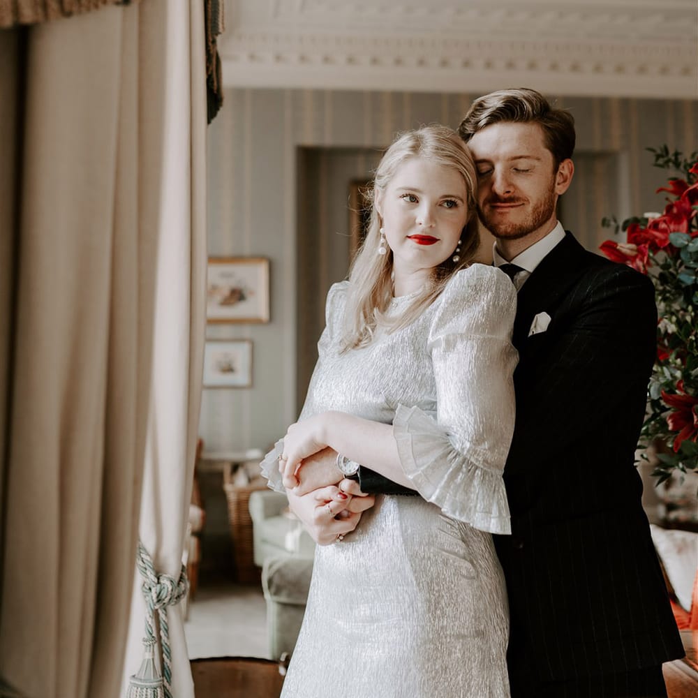 Groom in pinstriped suit embracing the bride from behind wearing metallic wedding dress with shoulder pads and ruffle sleeves 