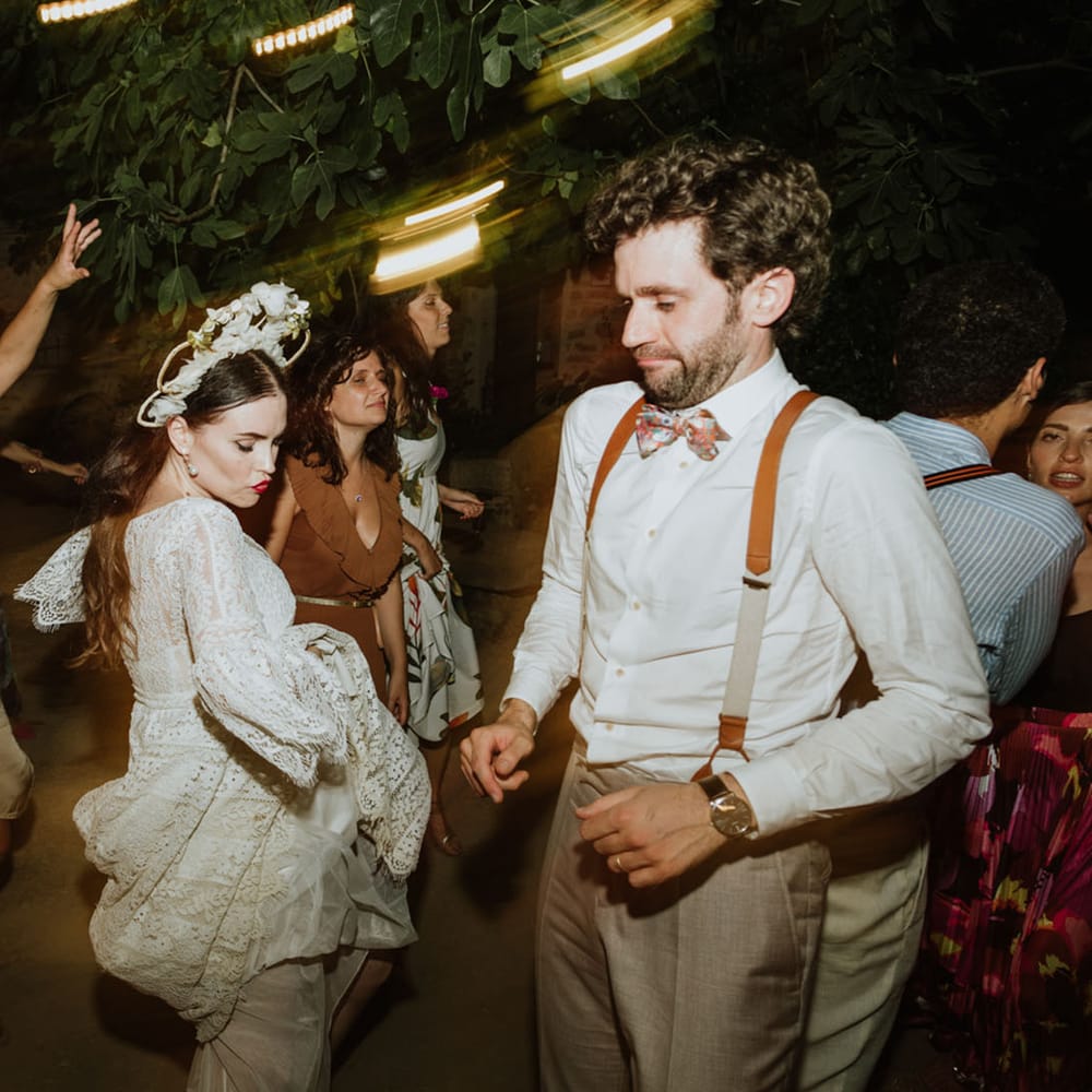 Bride in lace wedding dress, pearl earrings and bridal crown dances at wedding reception in Italy with groom in burnt orange braces and hwite wedding shirt