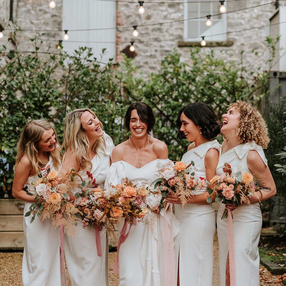 Bride in off the shoulder satin wedding dress standing with bridal party in courtyard wearing one shoulder white bridesmaid dresses 