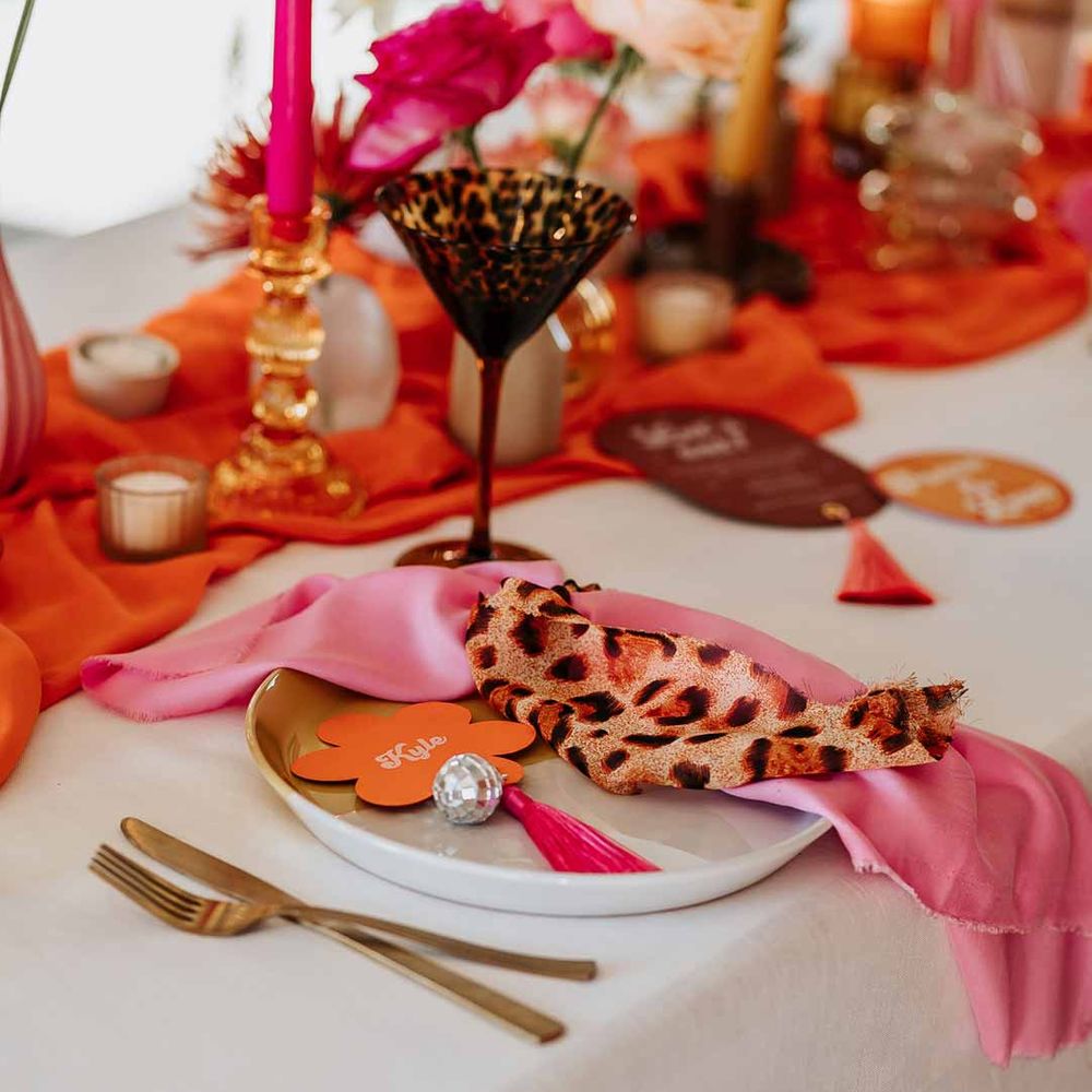 Leopard print wedding fabric fabric on top of pink wedding napkin on retro wedding tablescape with pink tapered candles, orange table runner, mismatched single flower vases, colourful flower centrepieces and large colourful flower arrangements 