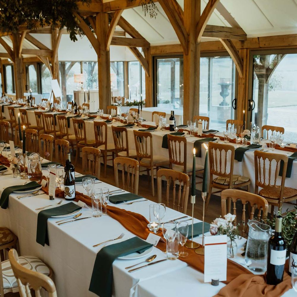 Winter themed classic wedding tablescape with white tablecloth, burnt orange wedding tablerunner, forest green wedding napkins, forest green tapered candles and bottles of wine with suspended foliage and fairy lights decor at wedding