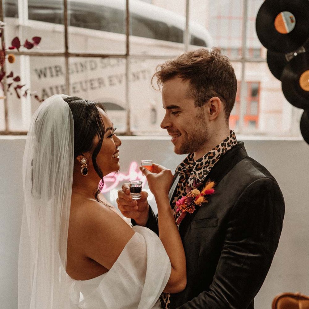 Groom in wet look black grooms suit with leopard print shirt and pink and orange boutonniere standing by vinyl wedding arch with bride in off shoulder tulle short wedding dress with church length veil doing wedding shots 