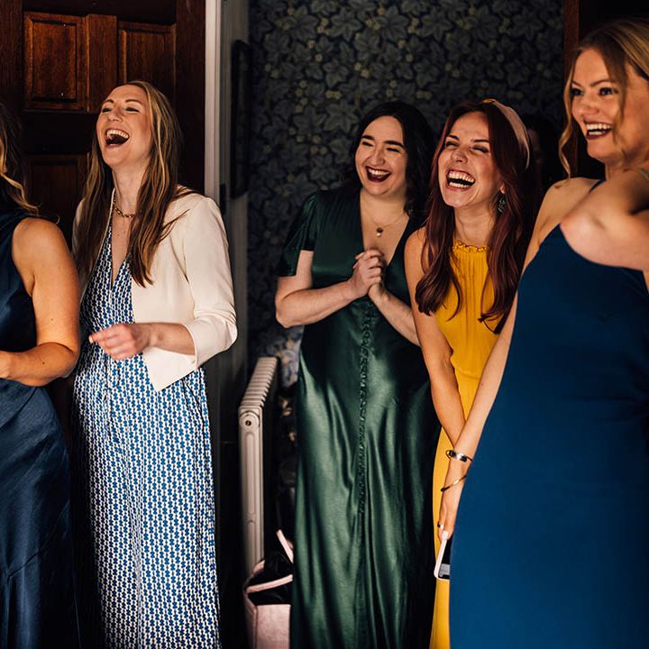Bridesmaids in mismatched green, blue, and yellow bridesmaid dresses getting their first look at the bride 