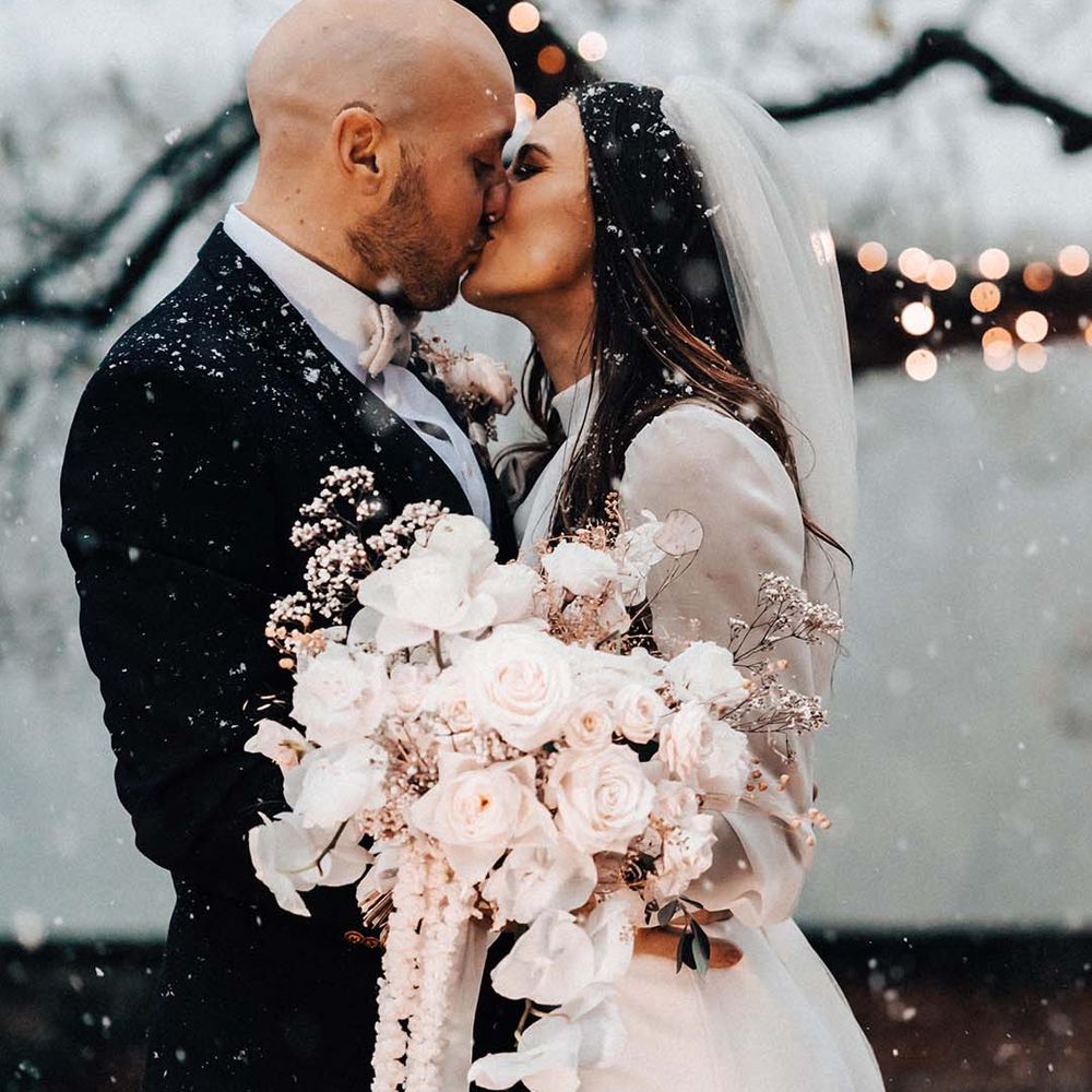 Bride wearing long sleeve wedding dress carrying white flower wedding bouquet kissing the groom as snow falls around them 