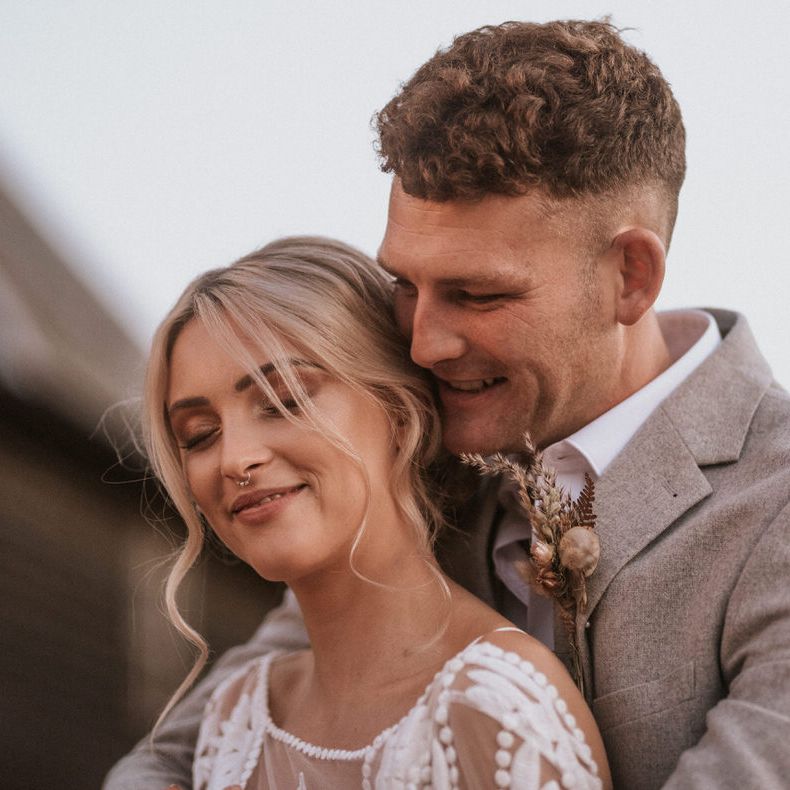 Bride and groom embrace, the groom has pampas grass detailing on his blazer lapel