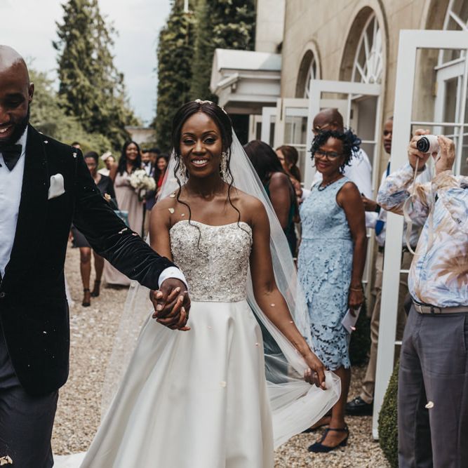Black groom in a velvet tuxedo and bow tie holding his brides hand in a strapless Oleg Cassini wedding dress during the confetti moment 