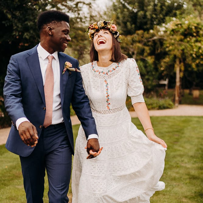 Groom in a navy suit with red patterned tie, smiling at his bride in a laser cut dress with colourful embroidery detail 