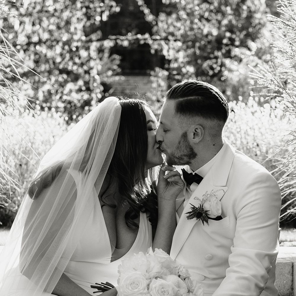 Bride in V neck Made With Love wedding dress and veil holding white rose bouquet sits outside with groom in white double breasted tuxedo jacket