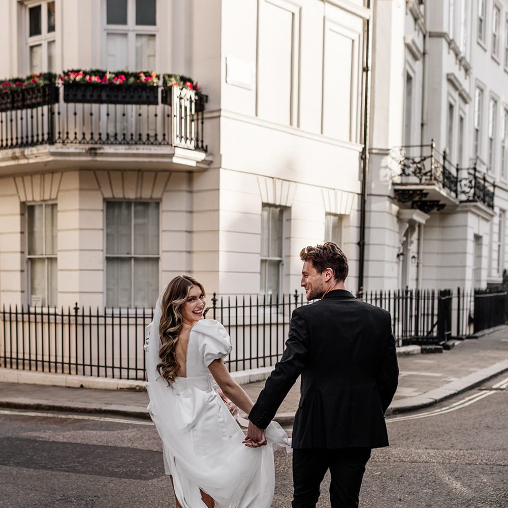 Bride with long wavy hair wearing a short wedding dress with open back holding hands with her husband in the streets of London 