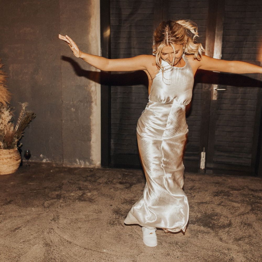 Bride with curled ponytail in silver satin dress and white air force trainers dances in the hut at Anran Devon for wedding evening party