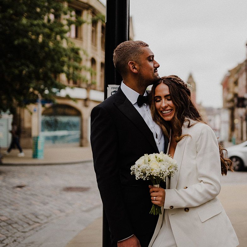 Bride in a white blazer holding a white rose and gypsophila bouquet snuggling into her groom in a black tuxedo 