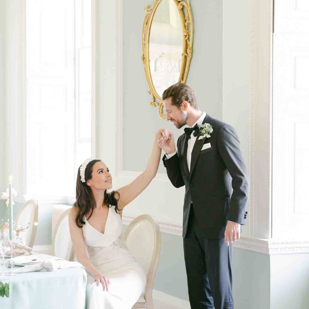 Groom in a tuxedo kissing his brides hand as she sits at the wedding reception table decorated with pastel decor and flowers 