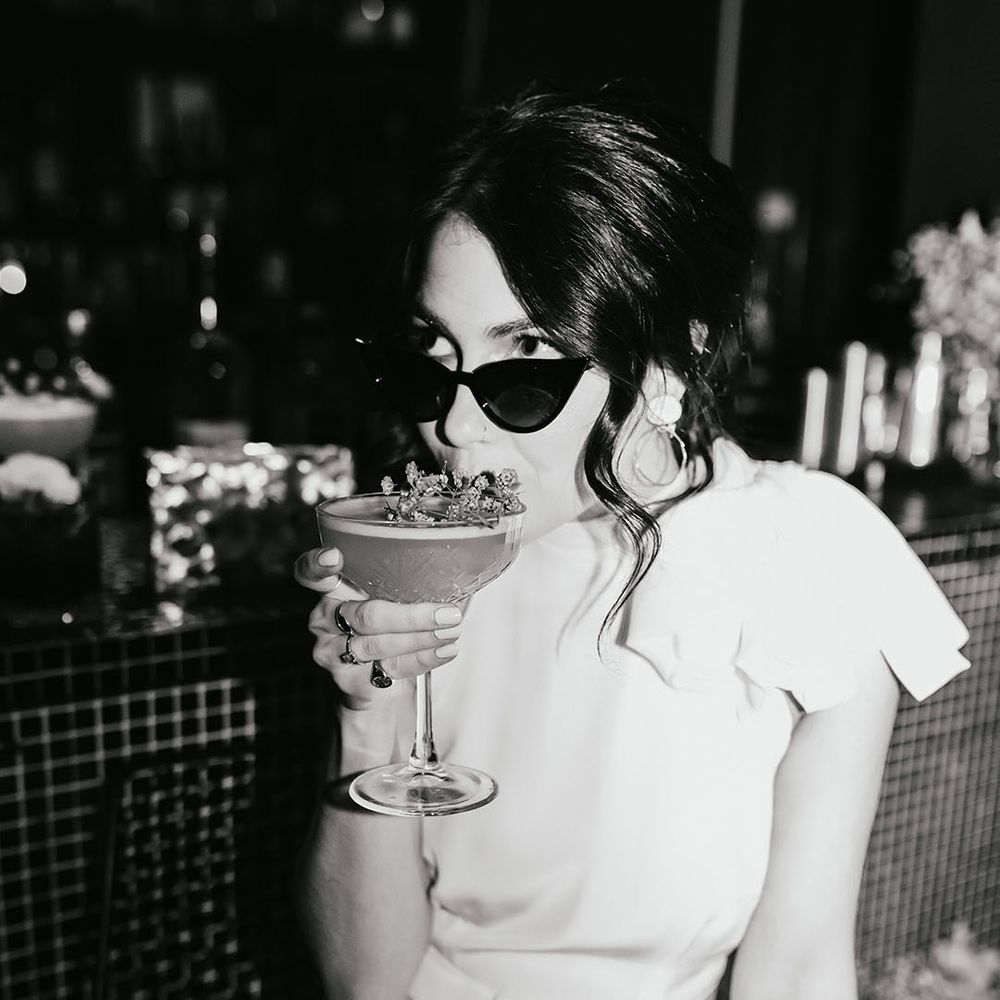 black and white picture of a stylish bride in a wedding dress with no sleeves wearing sunglasses and sipping cocktails on the bar 