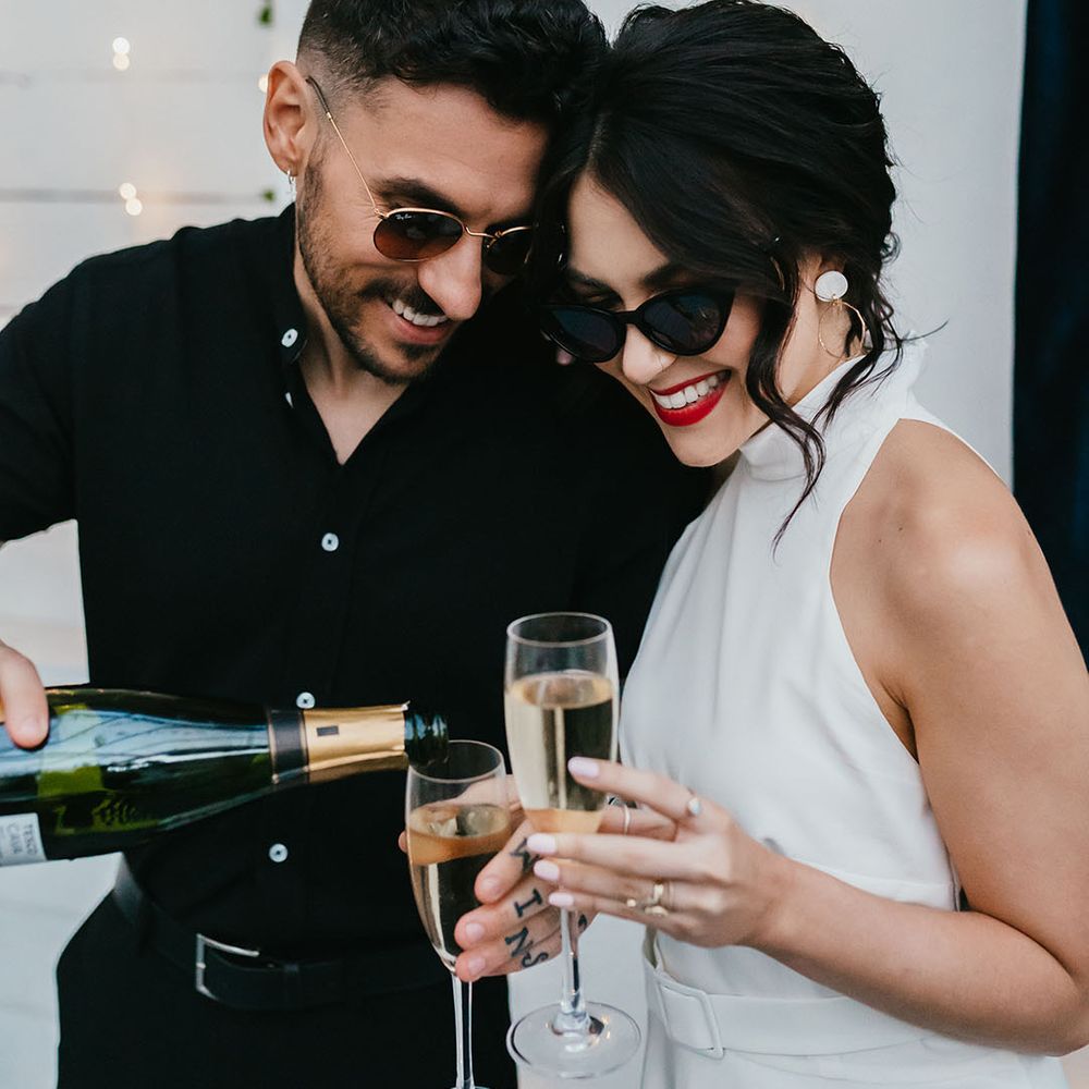 Groom in a black short sleeve shirt pouring champagne for his bride in a halterneck wedding dress with pinned up hair, sunglasses and red lipstick 