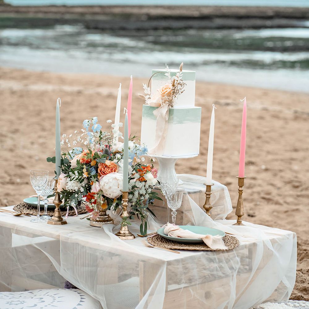 Wooden palette tablescape and cushions on the beach with pastel coloured candles and square buttercream wedding cake 