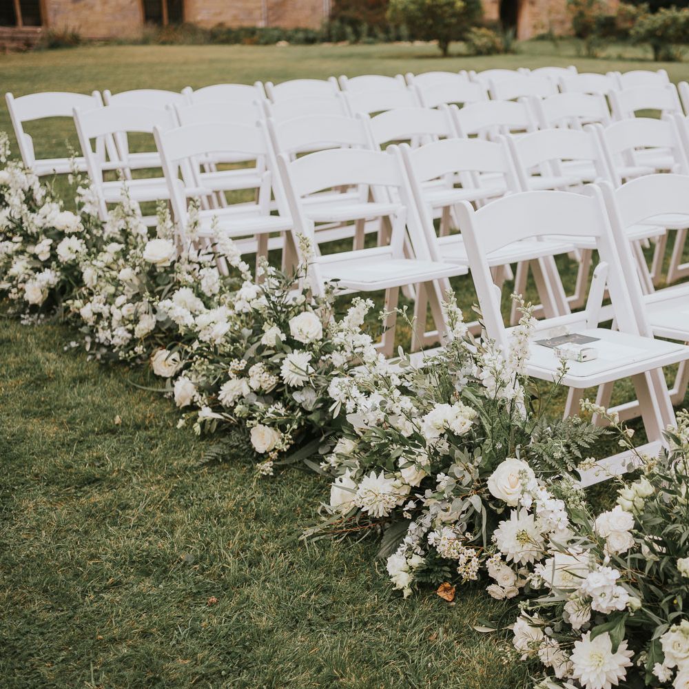 White wooden chairs with green and white floral lined aisle for outdoor wedding ceremony in Buckinghamshire