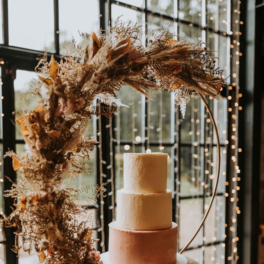 Three tier wedding cake with white and pink icing on large boho hoop cake stand with dried floral decor for rustic barn wedding