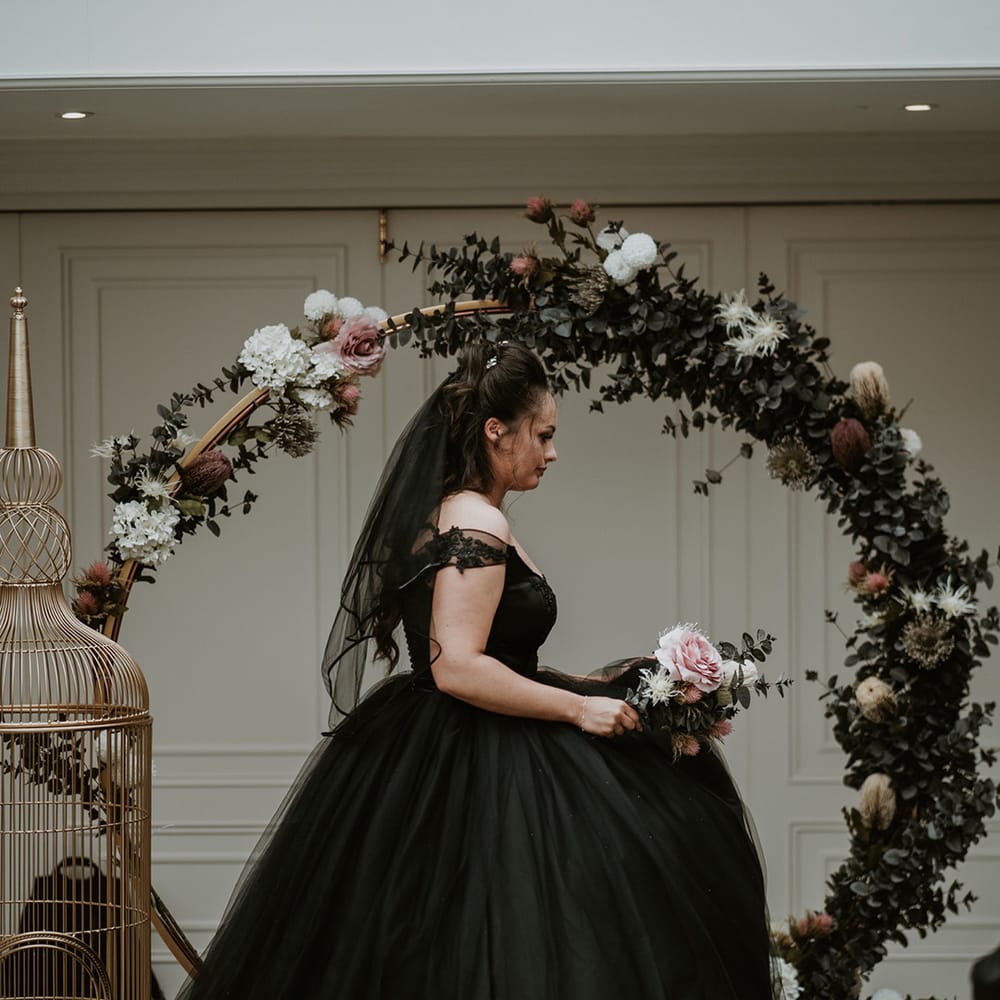 Bride wears black wedding dress whilst standing in front of floral archway complete with pastel florals 