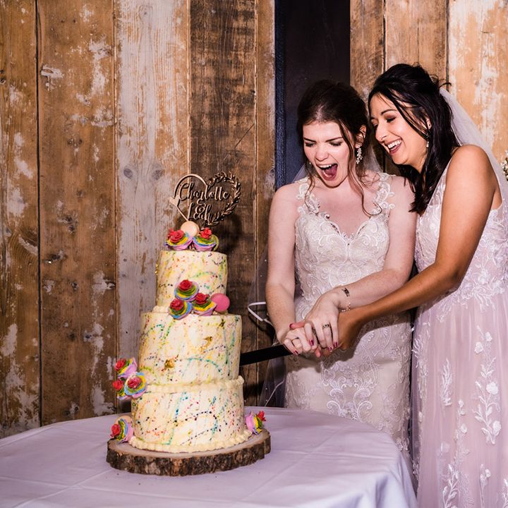 Brides cut their white frosted rainbow paint splattered wedding cake with rainbow iced roses, macarons and personalised wooden heart topper