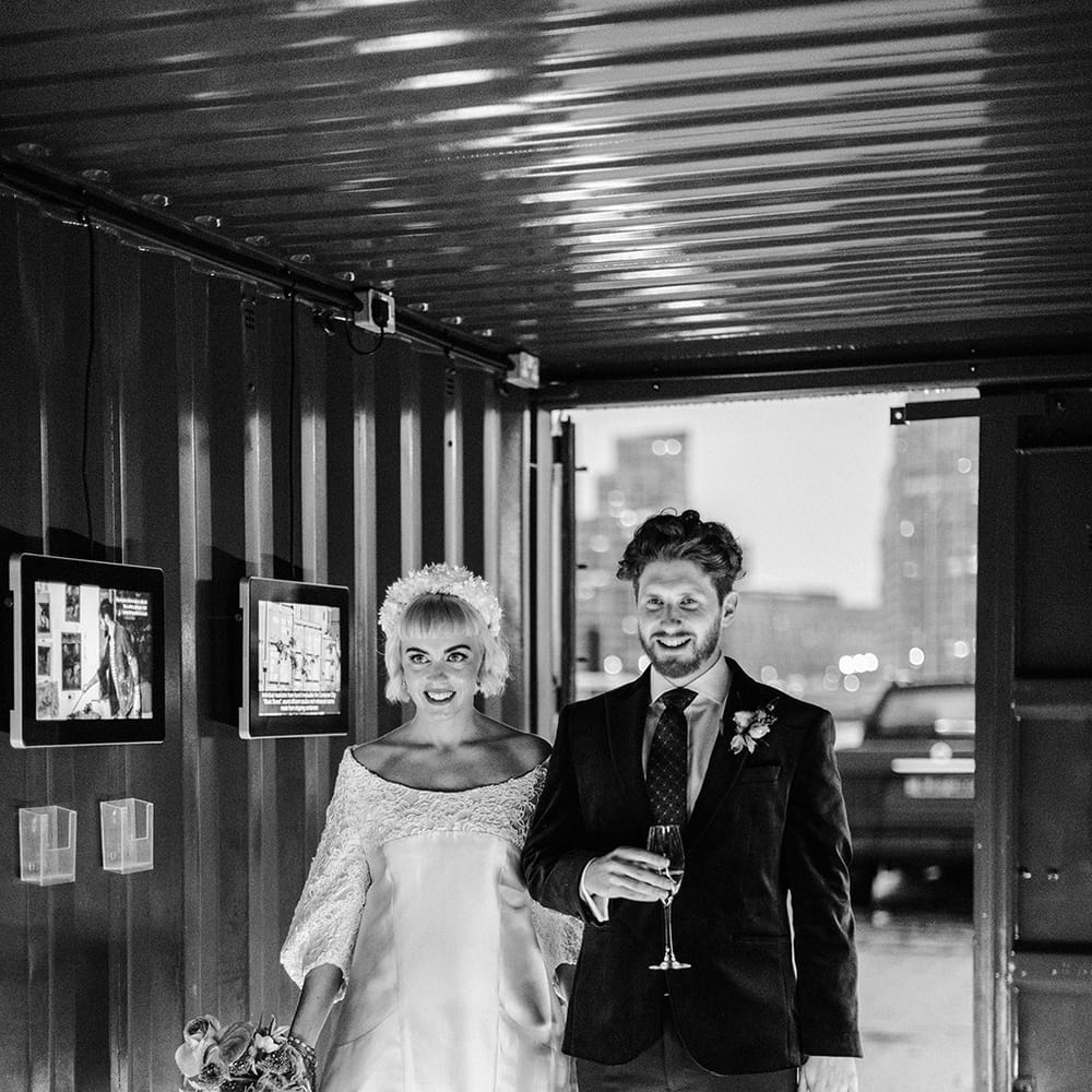 Bride in short wedding dress holding a bouquet walks into reception with groom holding glass of champagne
