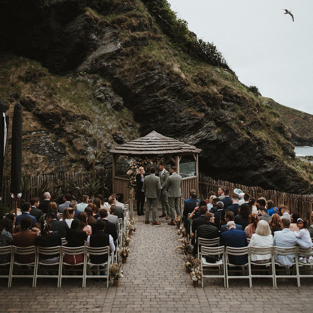 Wedding guests sit outdoors at Tunnels Beach in Devon as florals line the aisle and beach hut sits to the front