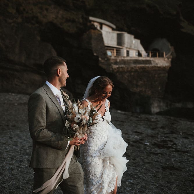 Groom holds champagne and rust coloured bridal bouquet as he walks along the beach with his bride for couples portraits 