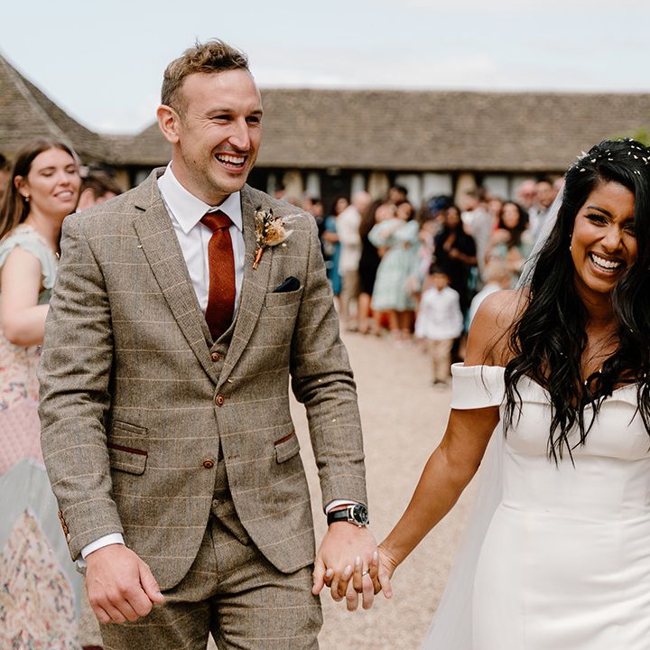South Asian bride in an off the shoulder wedding dress with the groom holding hands in a brown suit and orange tie 