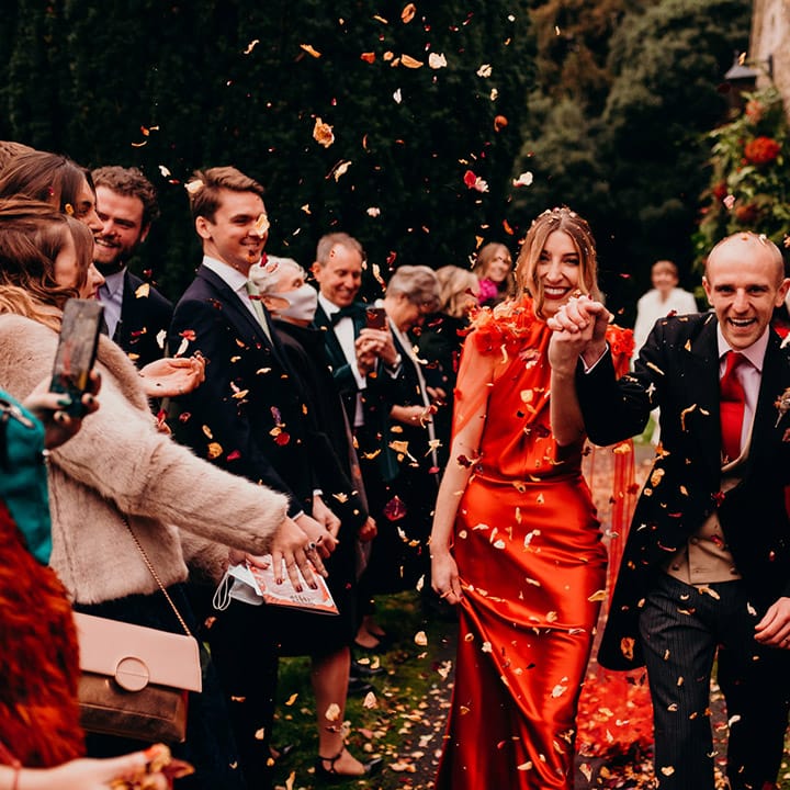 Bride in satin red wedding dress with cape and groom in three piece suit with red tie doing confetti walk at their winter wedding