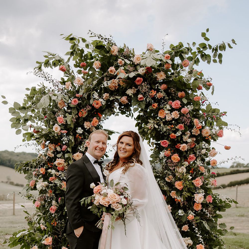 Bride in sheer full sleeved wedding dress stands with her groom in front of orange floral archway outdoors at Botley Hill Barn