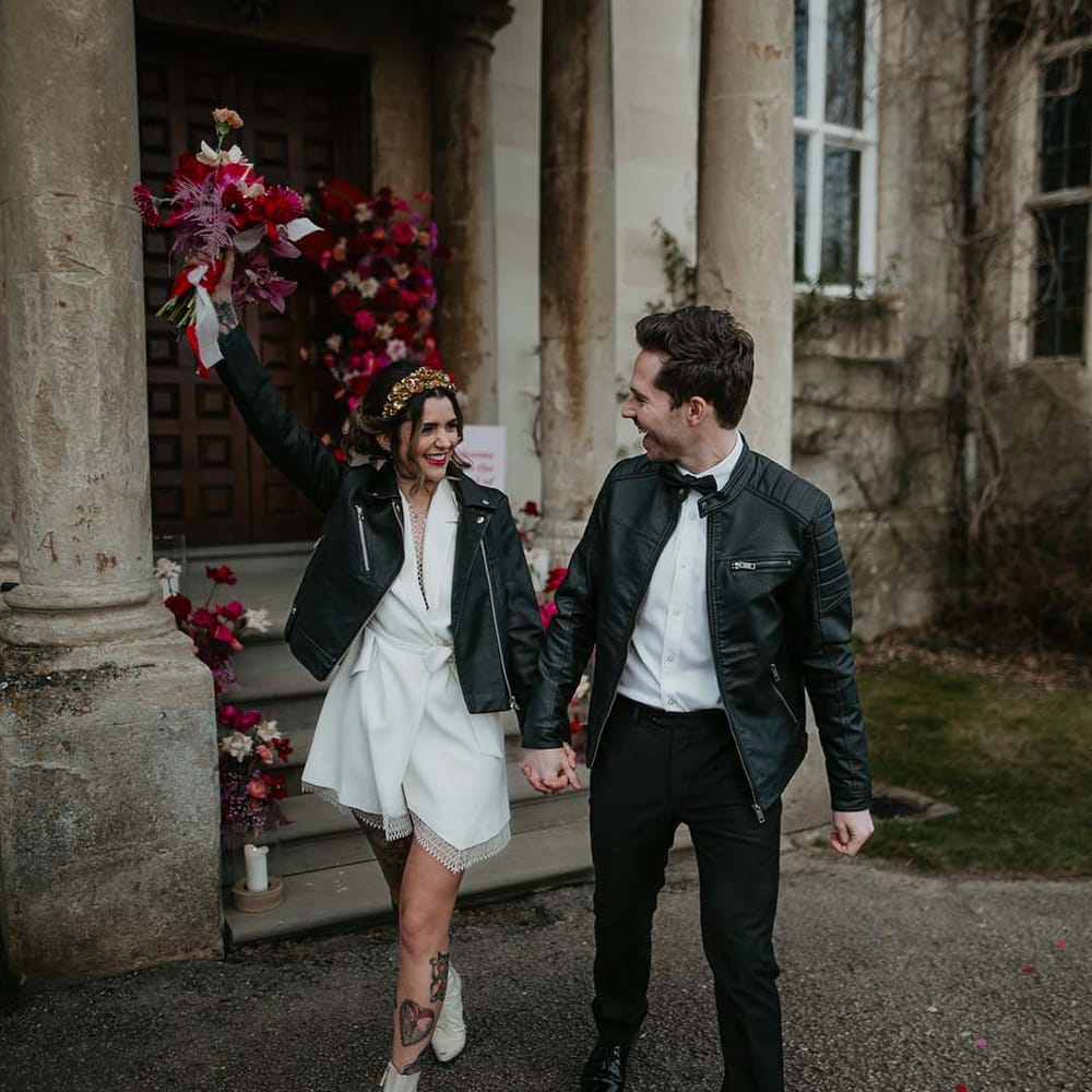 Bride in  blazer style wrap around short wedding dress with delicate fringe detailing and personalised, embroidered black leather jacket holding hot pink, bright pink and viva magenta bridal bouquet with dahlias, orchids, roses, amaryllis, anthuriums and dried flowers and groom in white shirt, black bowtie, black suit trousers and slim fitting black leather jacket 