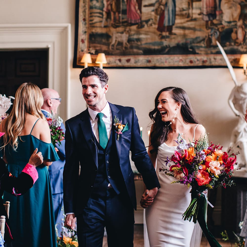 Bride with pearl earrings and satin wedding dress walks back down the aisle with the groom in a navy suit as a married couple 