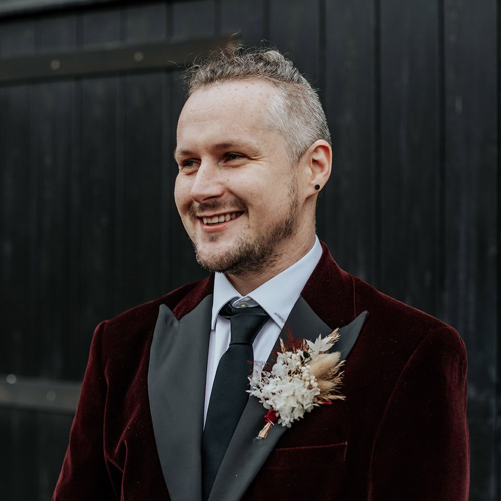 The groom wears a burgundy velvet suit jacket with a dried flower buttonhole and a black tie 