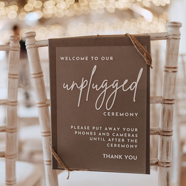Grey and white unplugged wedding ceremony sign tied to the back of the white wooden chairs for the rustic wedding 