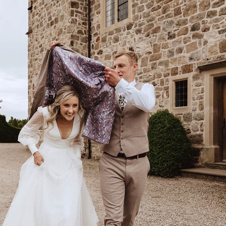 The groom puts his beige suit jacket over the bride's head to protect her from the rain on their wedding day 