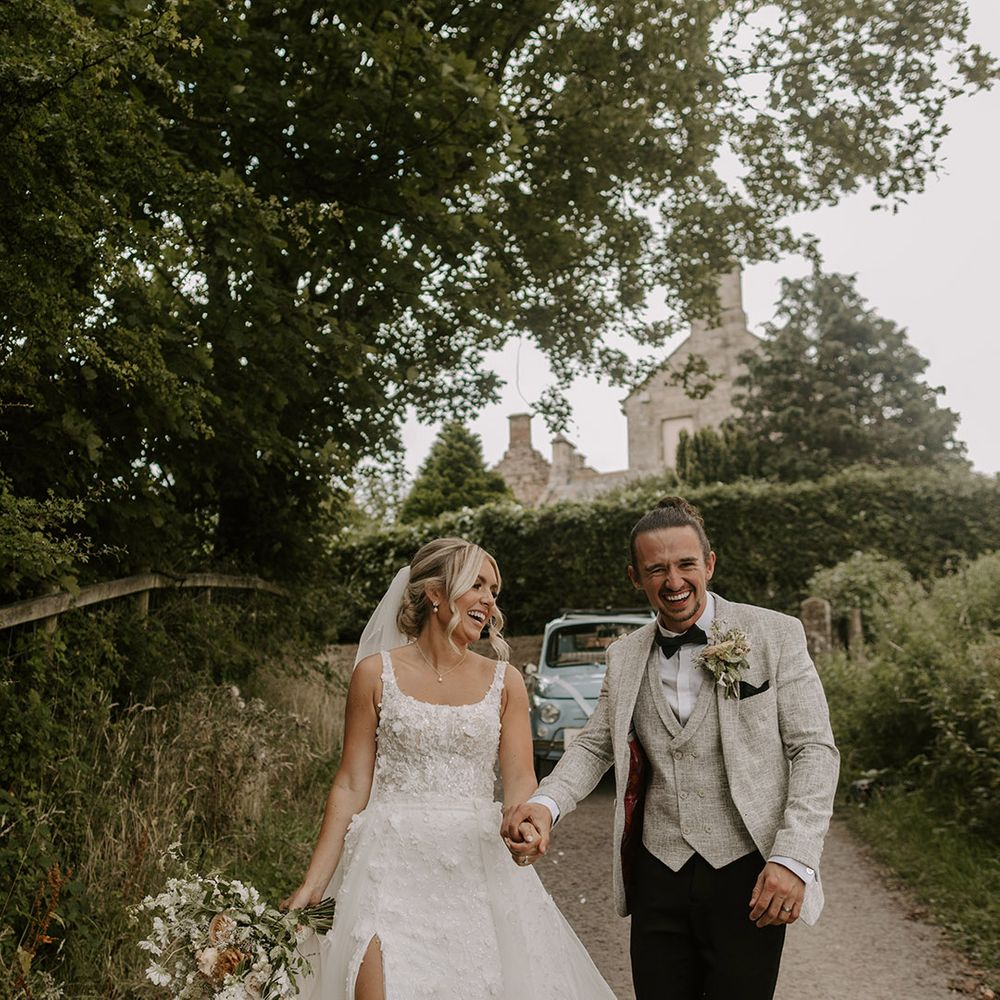 Bride in Eleganza Sposa Iconic wedding dress with detachable skirt and split leg laughing with the groom in light grey three piece suit 