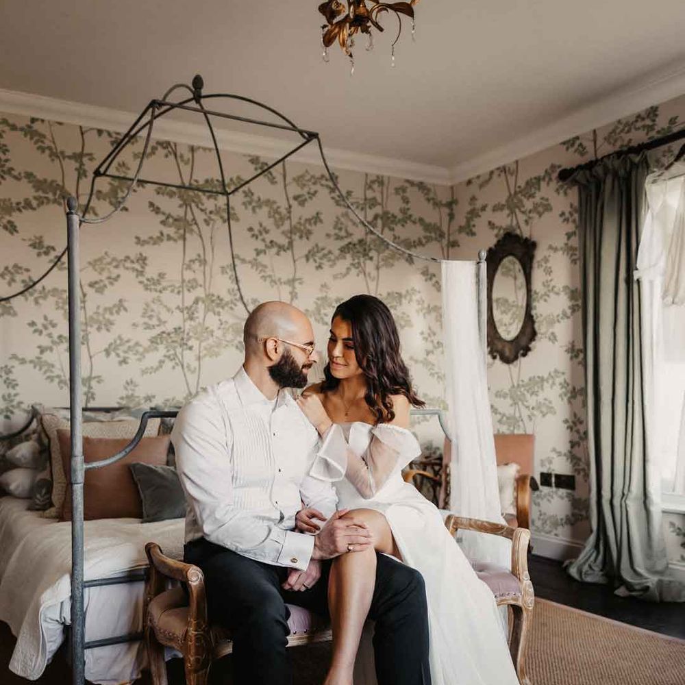 Bride in sheer puff sleeve wedding dress with front slit sitting with groom in white shirt and black suit trousers 