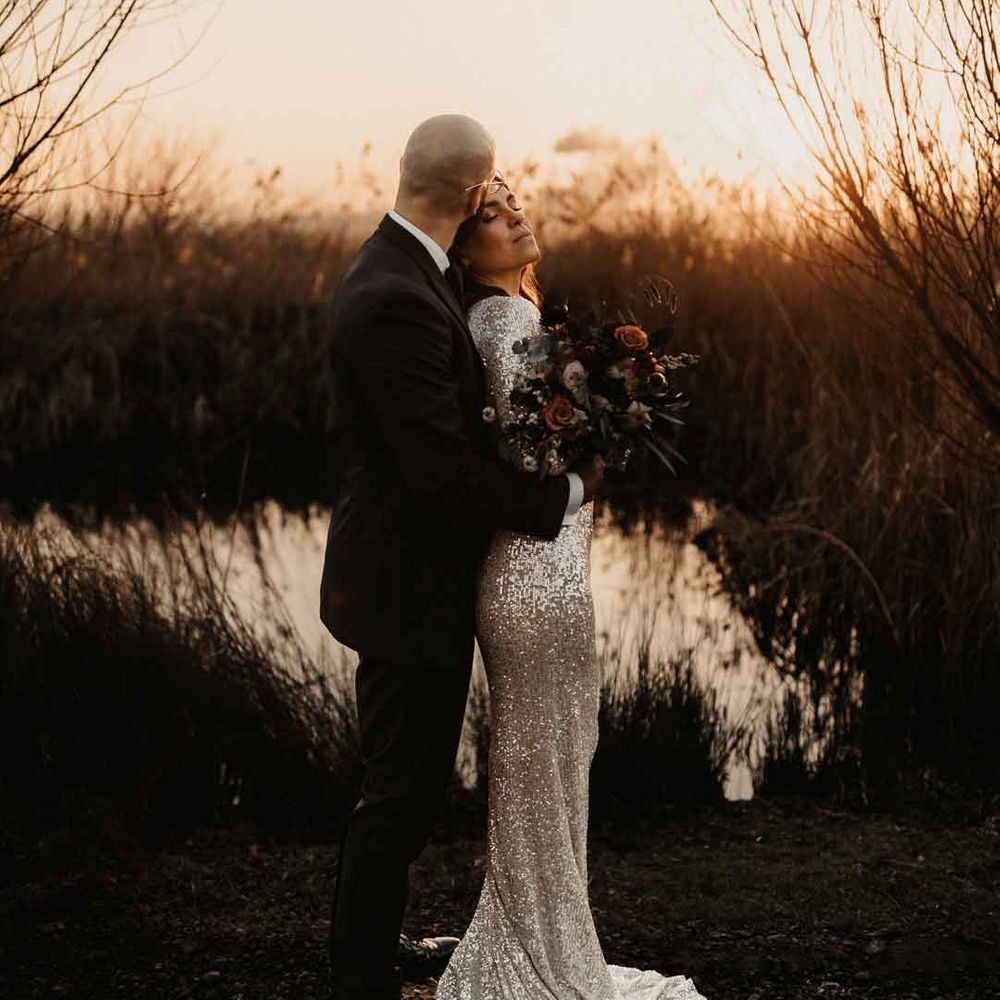 Bride in silver sparkly wedding dress embracing groom in classic black tuxedo doing golden hour couples portraits at Elmley Nature Reserve wedding venue 