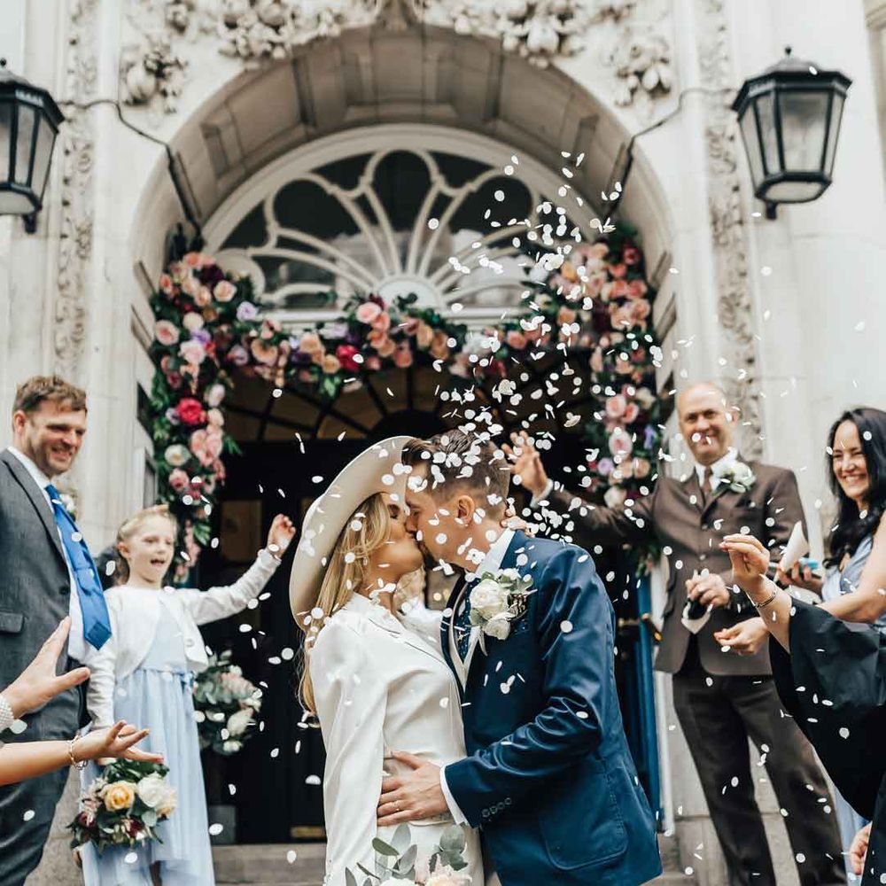 Bride in long sleeve white satin suit set and bridal hat kissing groom in navy blue suit at small intimate wedding 