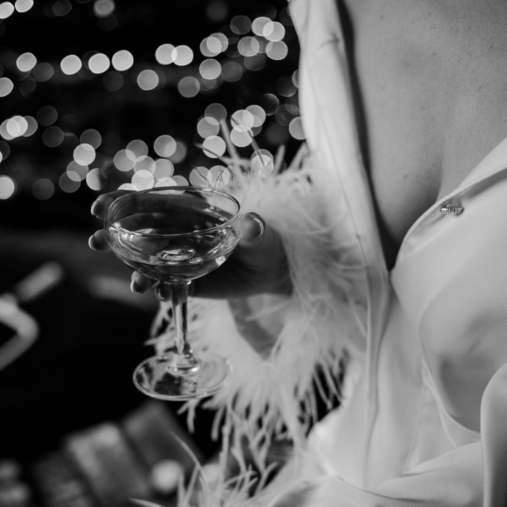 Bride drinks from champagne coupe glass in her white satin pyjamas with feather cuff detail for the wedding 