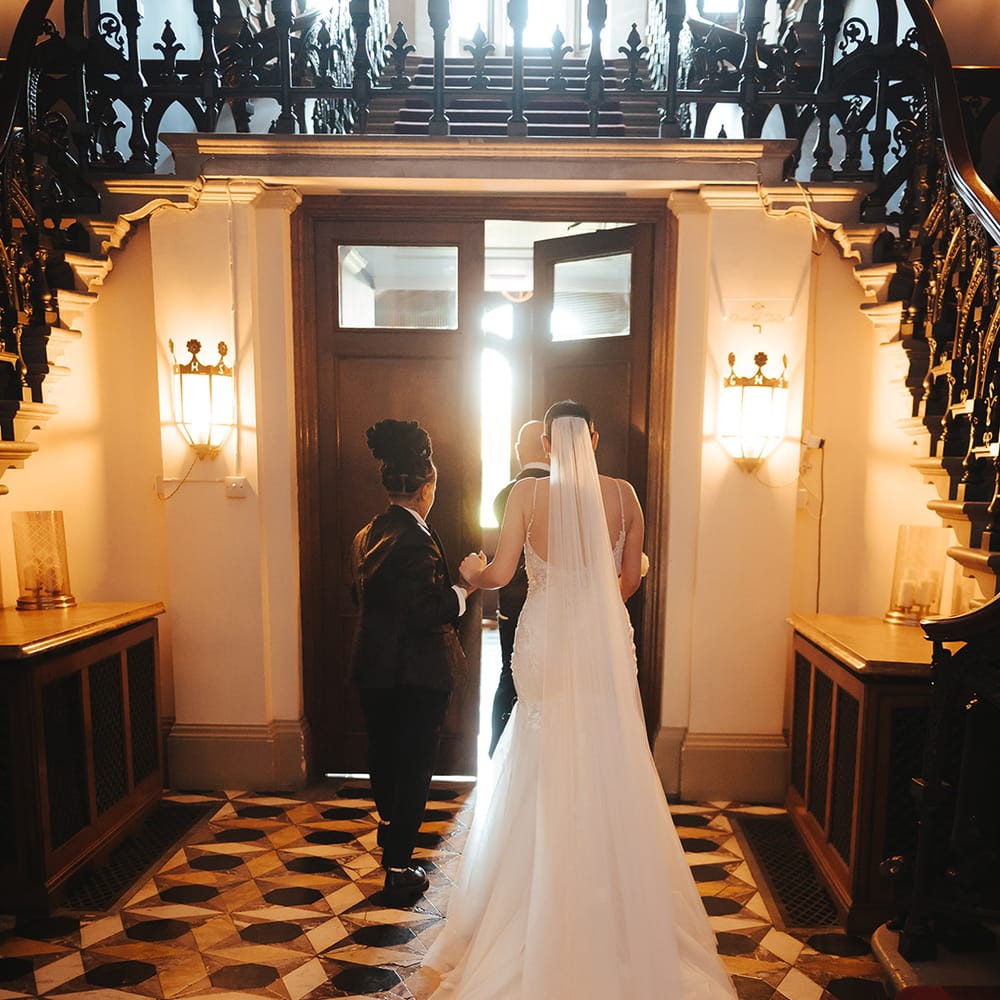 Bride in lace sleeveless v neck Enzoani wedding dress with tulle finish and church length embroidered lace 3d appliqué veil holding hands with bride in black tie tuxedo at Grittleton House